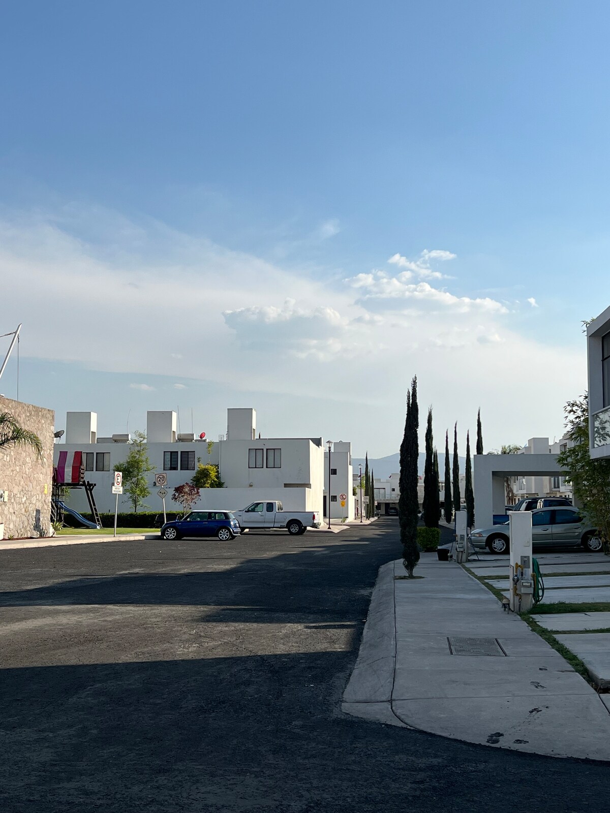 A serene residential street features modern white buildings lined with tall cypress trees. The image captures a clear blue sky and soft clouds, enhancing the peaceful ambiance of the neighborhood.