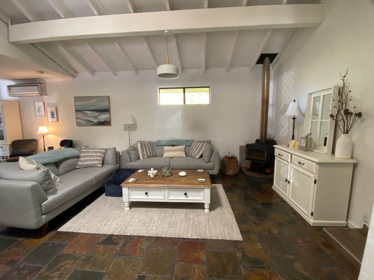 A spacious living area is highlighted by a large sectional sofa and natural wood coffee table. The room features a stone floor, a cozy pot belly stove, and large windows allowing daylight to illuminate the space. Soft decorative elements enhance the inviting atmosphere.