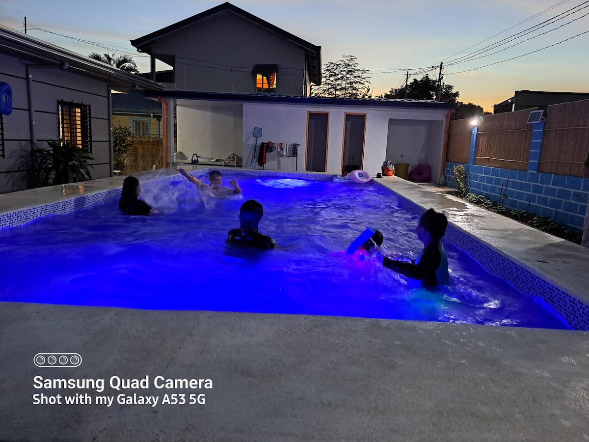 A shared swimming pool is illuminated with soft blue lights, creating a relaxed atmosphere as children enjoy water play. The surrounding area features simple outdoor furniture, with a calm evening sky visible in the background, enhancing the peaceful setting.