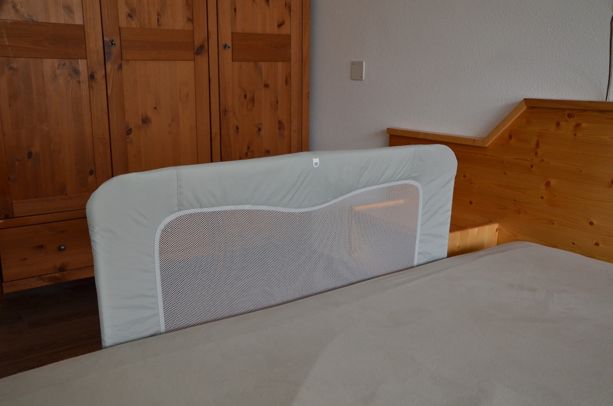 A spacious bed is positioned against a wooden wall, featuring a safety rail designed for added security. Natural light may filter through a nearby window, while wooden cabinets provide storage options alongside the bed.