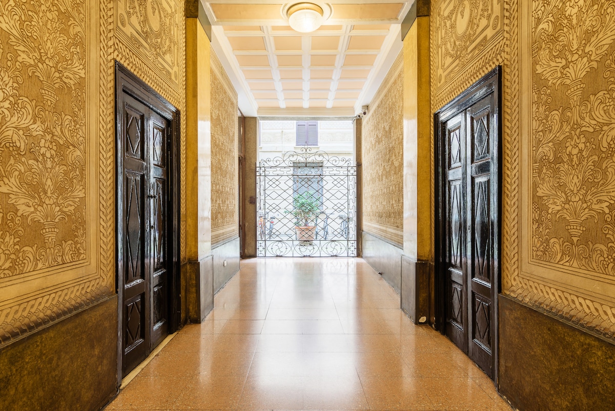 An elegant entrance hall is displayed, featuring decorative walls with intricate patterns and a polished floor. Two wooden doors are positioned symmetrically at the sides, leading to an intricately designed wrought iron gate that allows natural light to illuminate the space.
