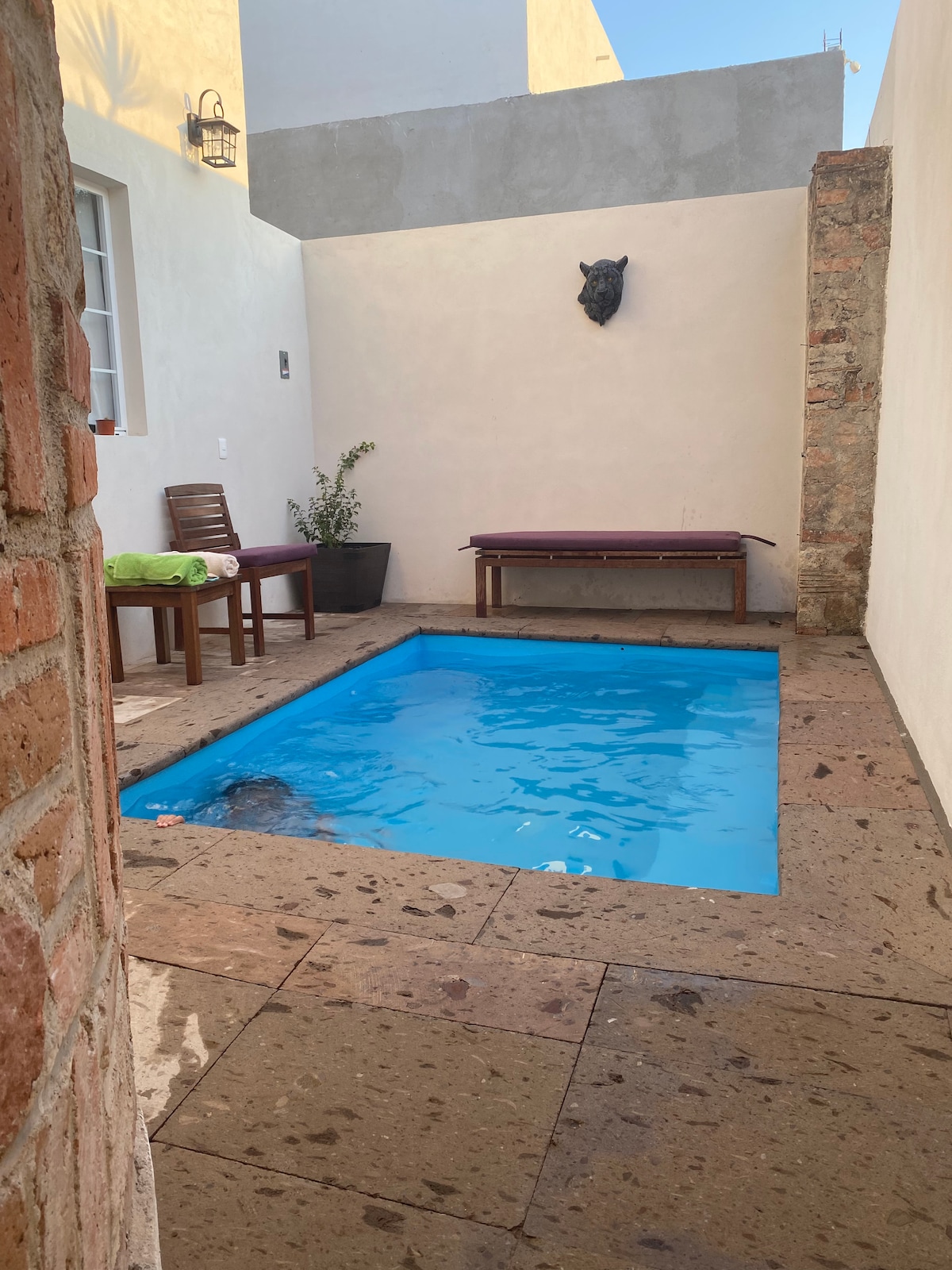 A private pool is set in a sunlit courtyard, surrounded by smooth stone walls. Two wooden chairs and a bench are positioned nearby, providing seating options. A decorative wall piece adds an artistic touch to the serene space.
