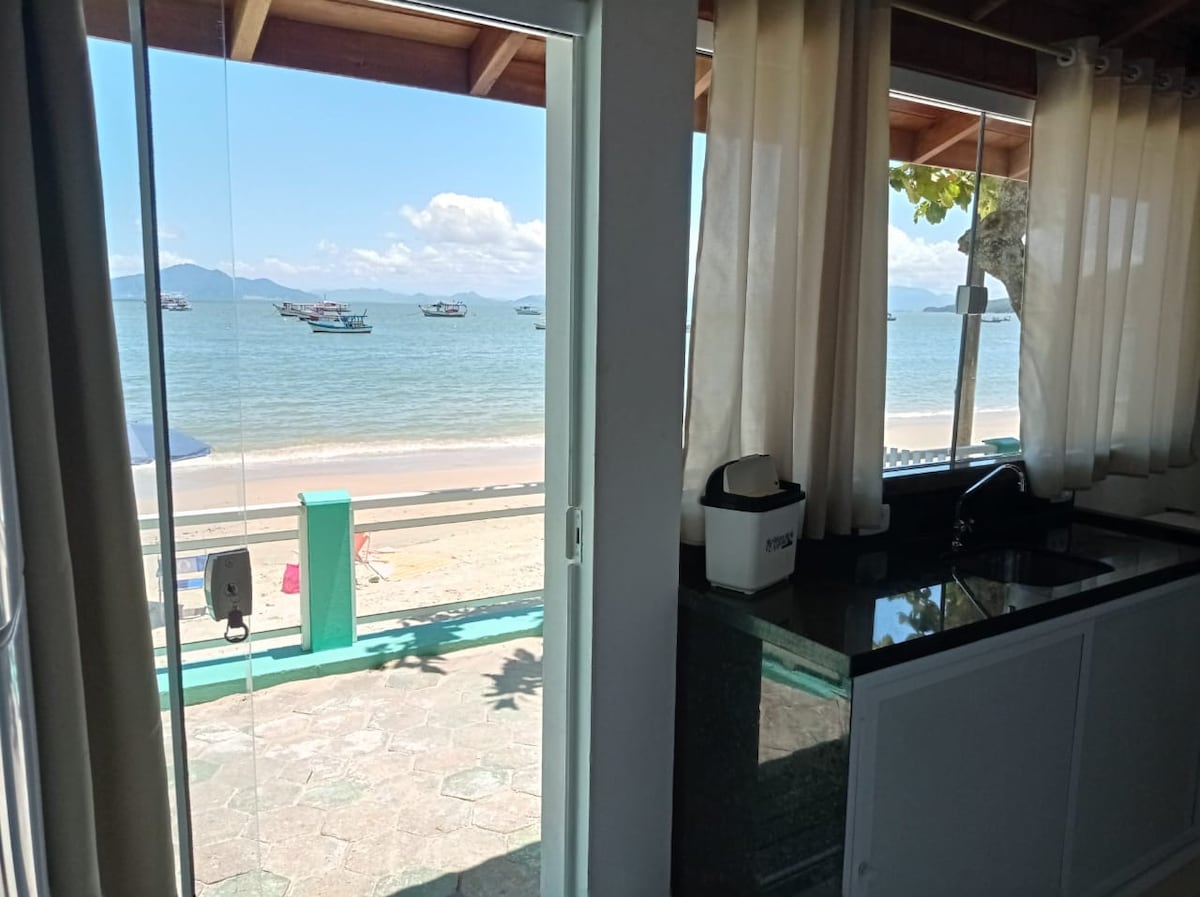 A view from the kitchen showcases large glass doors that open to a sandy beach and calm sea. Boats are visible in the distance under a clear sky, while light beige curtains softly frame the entrance, enhancing the connection between indoor and outdoor spaces.