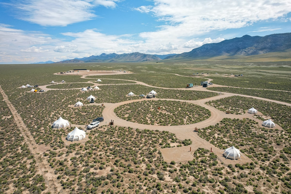 An aerial view showcases the expansive, grassy terrain of the campsite, with several round tents arranged along winding gravel paths. Surrounding mountains are visible in the distance, under a bright blue sky with scattered clouds.