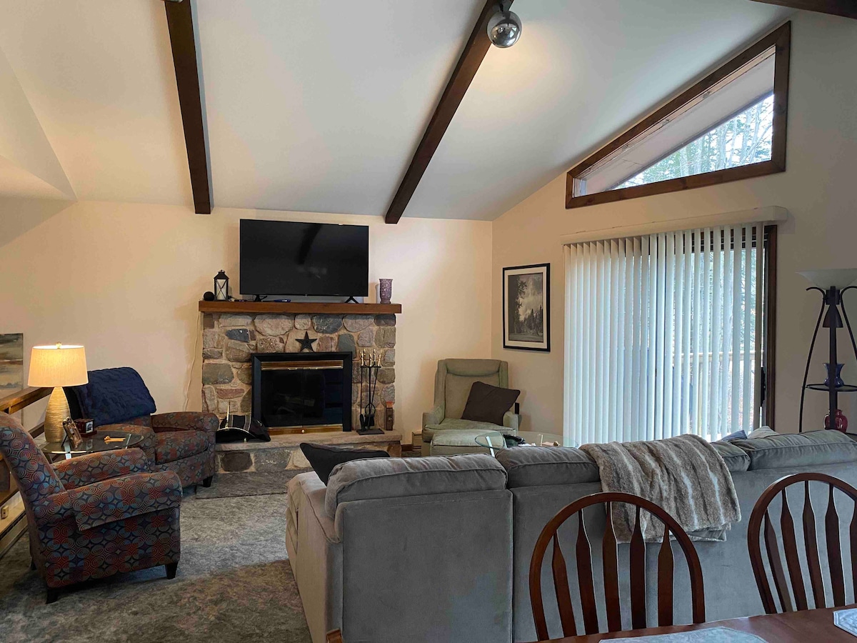 A cozy living area showcases a stone fireplace, flanked by comfortable seating arrangements. A television is mounted above the fireplace, with natural light filtering through large windows. Wood beams highlight the vaulted ceiling, creating a spacious atmosphere. Vertical blinds offer privacy when needed.