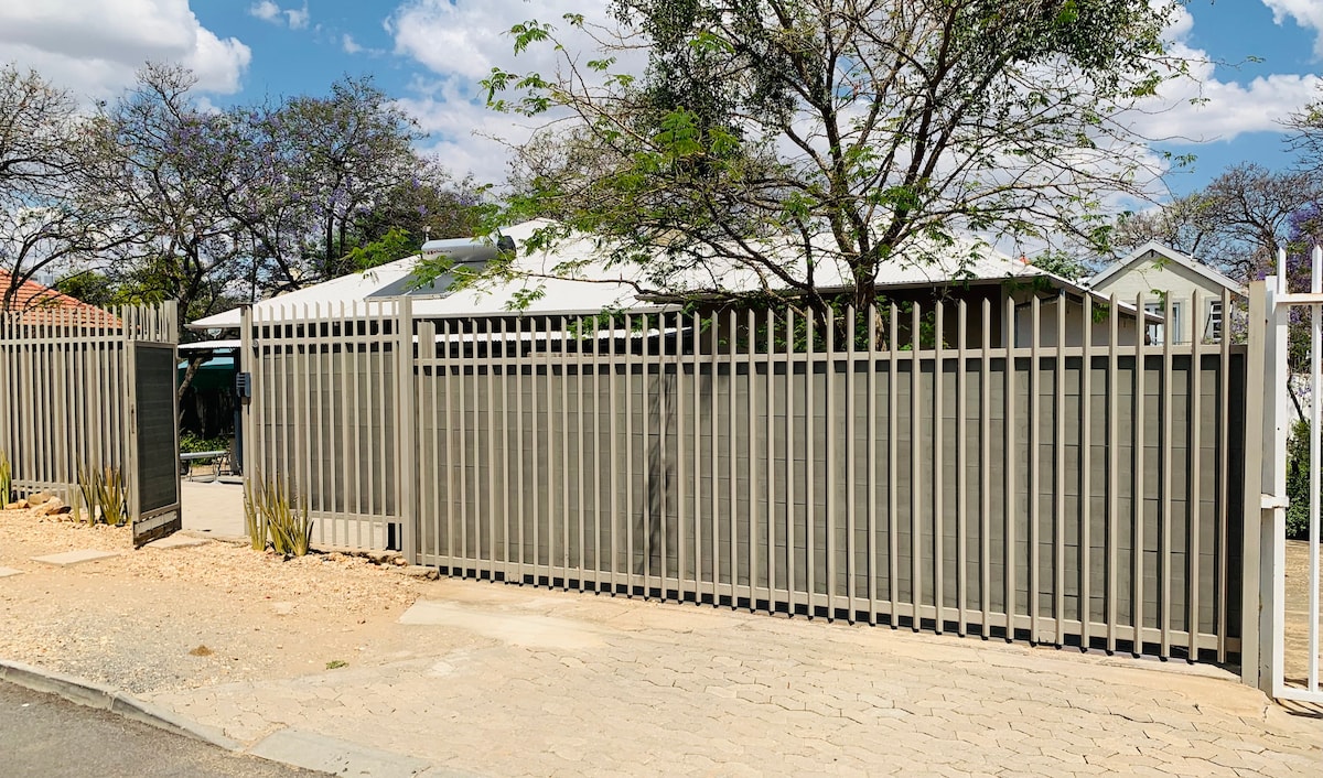 A secure, freestanding garden flat is enclosed by a tall wooden fence, offering privacy. The entrance features a gate that opens to a gravel driveway. Lush trees are visible beyond the fence, enhancing the natural surroundings.