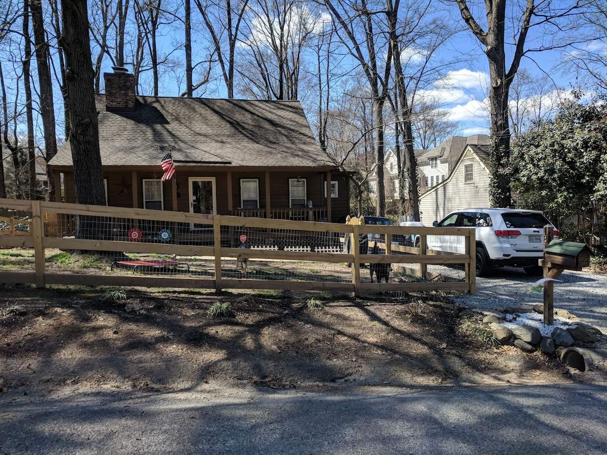 A log cabin-style home is set against a backdrop of tall trees. A wooden fence surrounds the front yard, which features a gravel driveway and a parked vehicle. The blue sky is dotted with clouds, adding to the serene outdoor atmosphere.