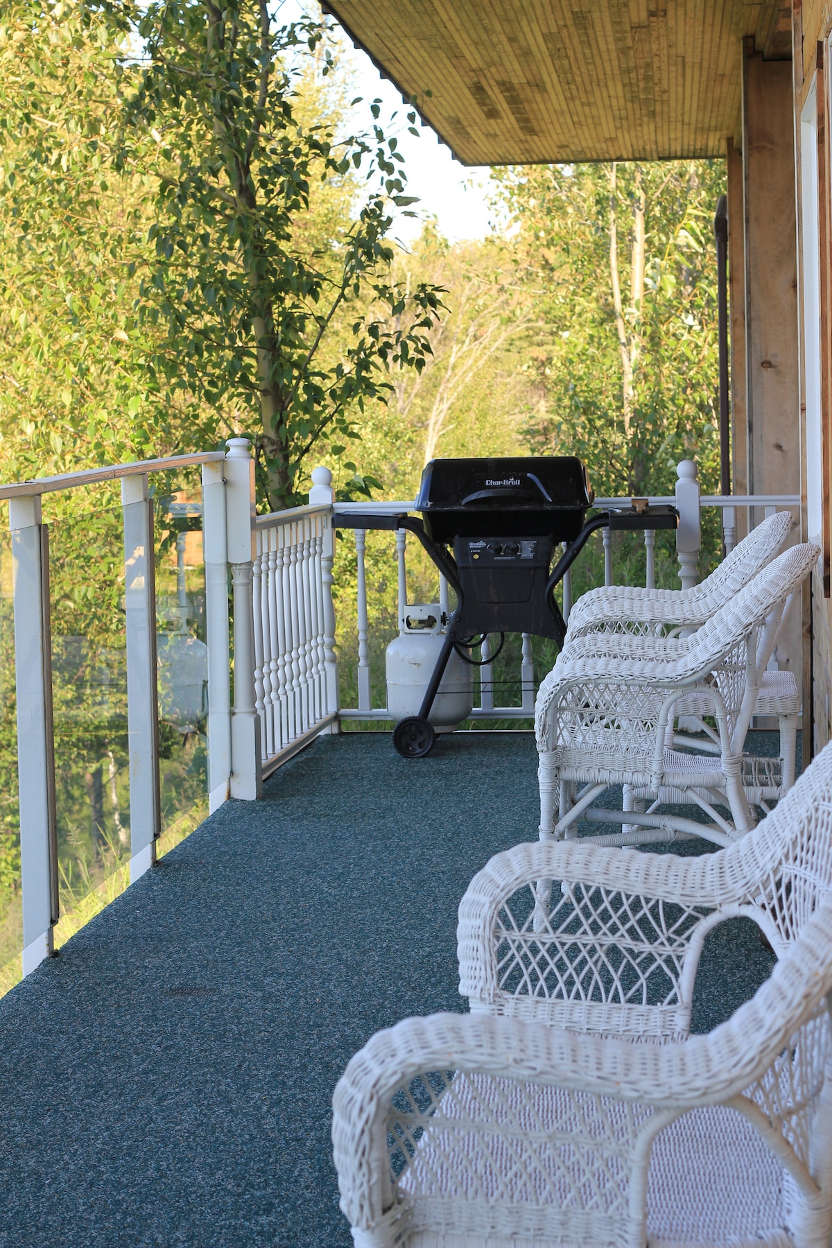 A covered balcony is shown with a green outdoor carpet, featuring two white wicker chairs positioned beside a black grill. Lush trees are visible in the background, providing a natural and serene environment.