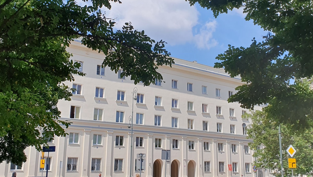 The building features a distinct 1950s socialist realist architecture with a white facade. Multiple windows are evenly spaced across the structure, allowing natural light to enhance its appearance. Lush green trees frame the scene, contributing to the inviting exterior.