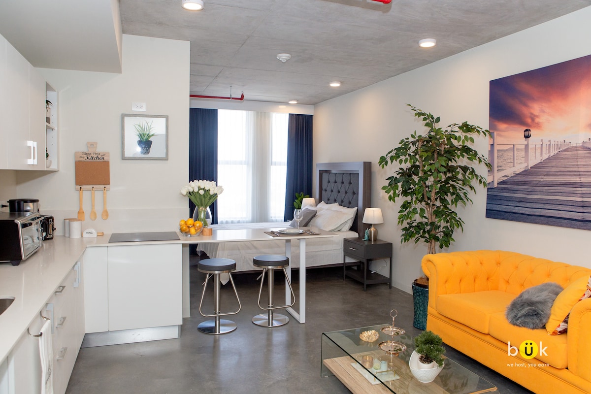 The open layout showcases a modern living space with a bright yellow couch, a glass coffee table, and a dining area. A queen bed is positioned to the rear, complemented by framed artwork and a potted plant, enhancing the tranquil atmosphere.