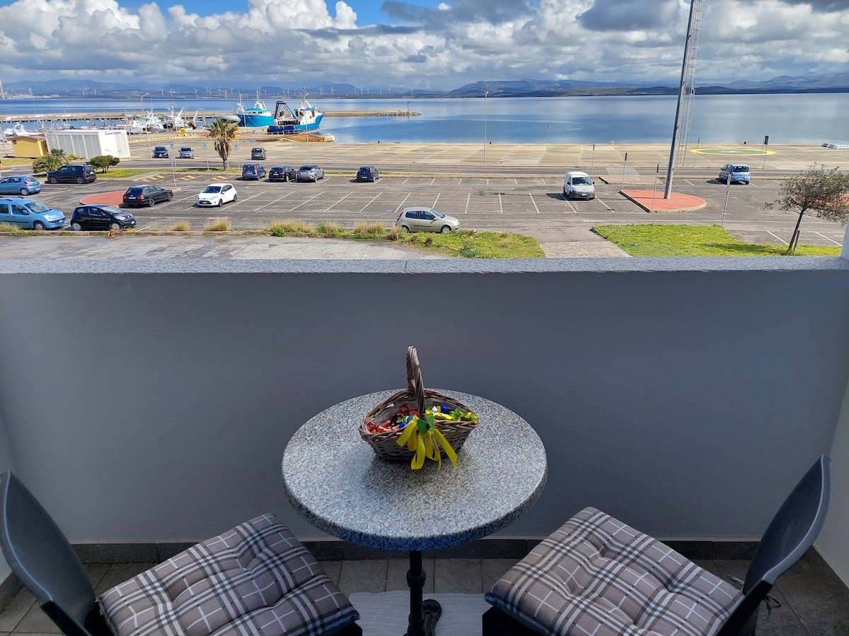 A small balcony features a round table with two cushioned chairs, overlooking a serene waterfront. A basket filled with fruit sits atop the table. Expansive views of the harbor and distant mountains can be seen under a partly cloudy sky.