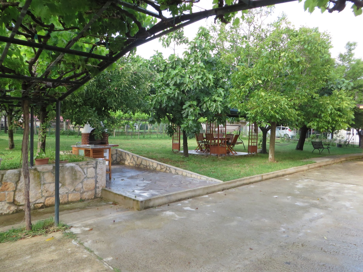 A spacious garden is displayed, featuring well-maintained grass and various trees providing shade. A wooden dining set is visible under a gazebo, accompanied by a stone barbecue area, creating an inviting space for outdoor dining and relaxation. Paved pathways connect different sections of the garden.