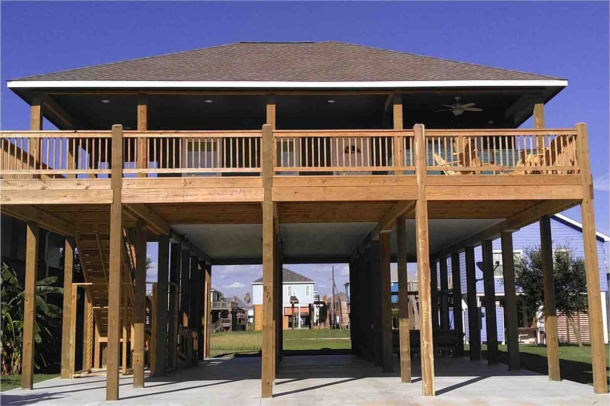 The house is elevated on stilts, featuring a spacious wooden deck with railings. A staircase leads up to the deck from the ground level. Large open areas underneath provide ample outdoor space for activities, surrounded by grassy landscaping.