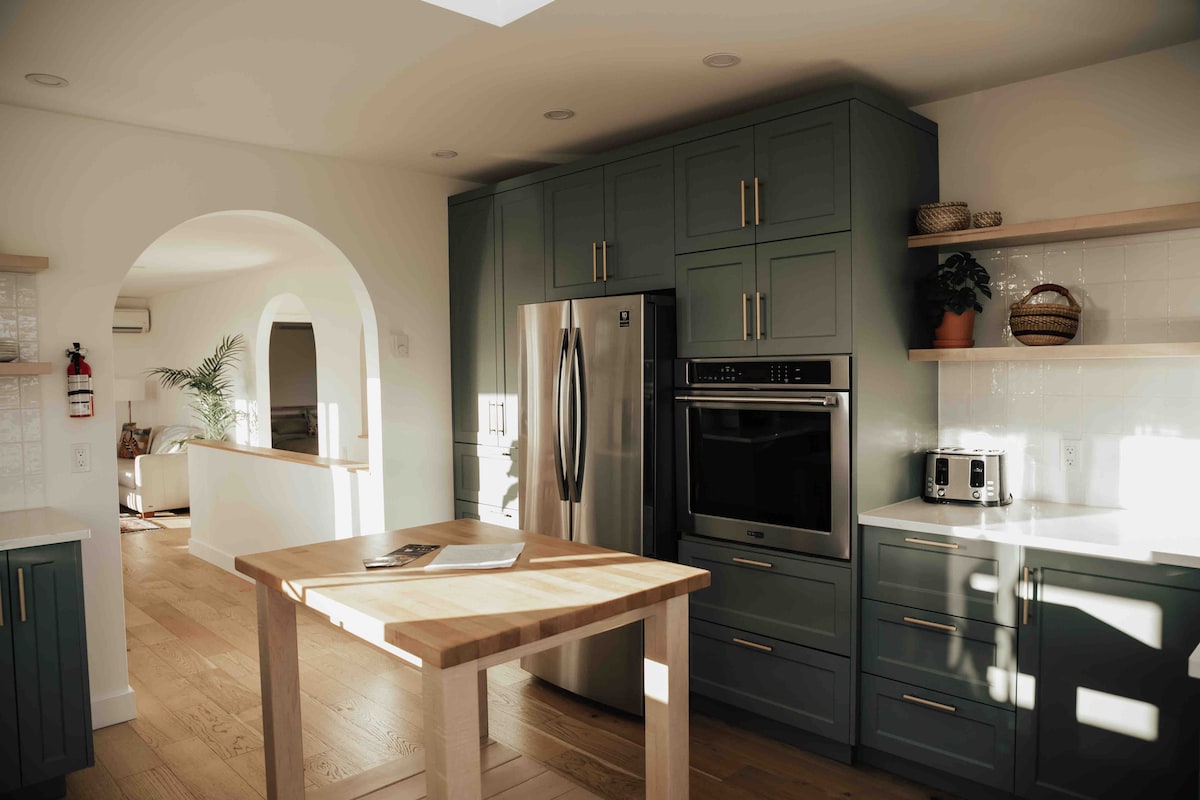 A modern kitchen is depicted, featuring green cabinetry and stainless steel appliances. A wooden island stands centrally, complemented by natural light streaming in through overhead windows. Open shelving displays decorative items, with a cozy living area visible in the background.