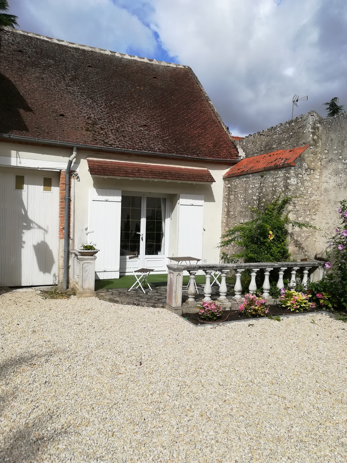 An entrance to the studio is framed by white shutters, leading to a small outdoor seating area with two chairs and a table. Lush flowers and gravel pathways contribute to a serene setting, while a stone railing adds architectural interest.