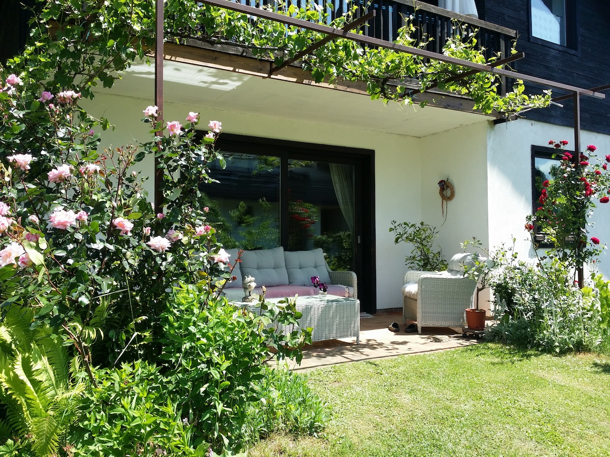 A charming outdoor seating area is presented with wicker furniture and soft cushions, situated under a shaded balcony. Green shrubs and blooming roses surround the space, enhancing the garden ambiance. Light filters through the large windows, inviting a connection between indoor and outdoor living.