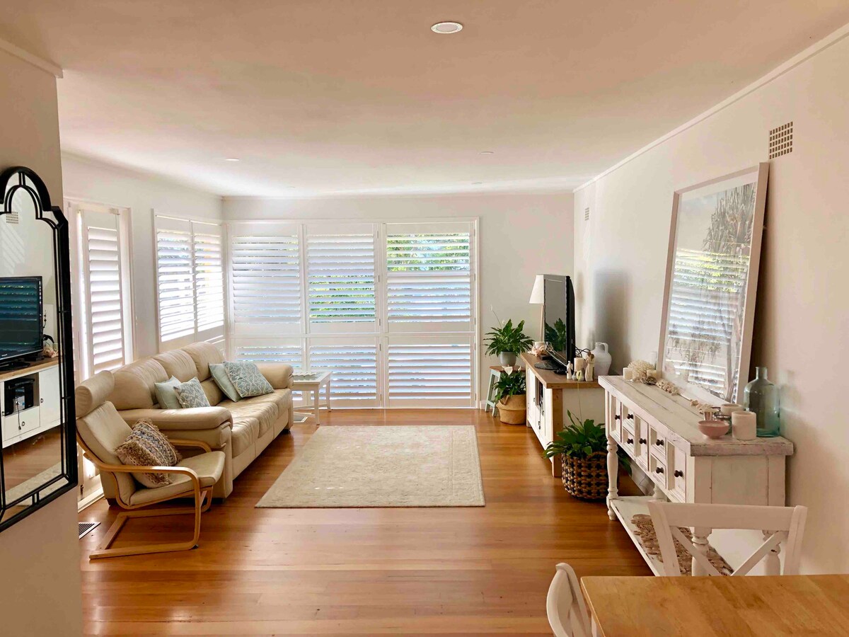 The living area features natural light streaming through plantation shutters, illuminating the new timber floorboards. A comfortable sofa is positioned alongside a stylish sideboard and a flat-screen TV, with green plants enhancing the inviting space. A neutral area rug provides warmth underfoot.