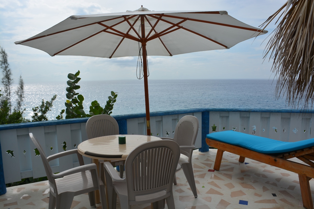 An outdoor terrace offers a round table surrounded by four light-colored plastic chairs, shaded by a large white umbrella. A wooden lounge chair provides additional seating, with a backdrop of the ocean visible beyond a low stone railing.