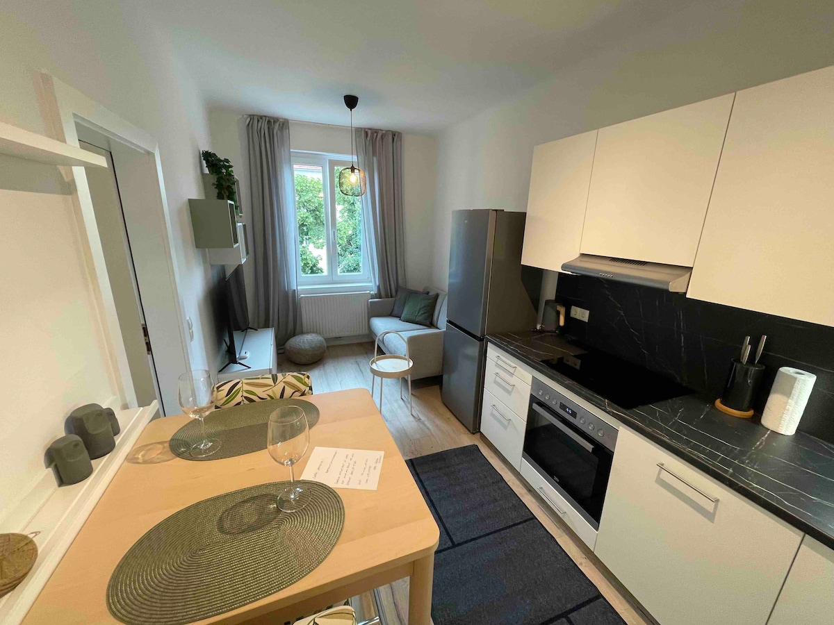 A modern studio kitchen is showcased with sleek cabinetry and a large refrigerator. A comfortable seating area with a light sofa is visible near a window, allowing natural light to fill the space. A dining table with placemats sets a cozy dining scene.