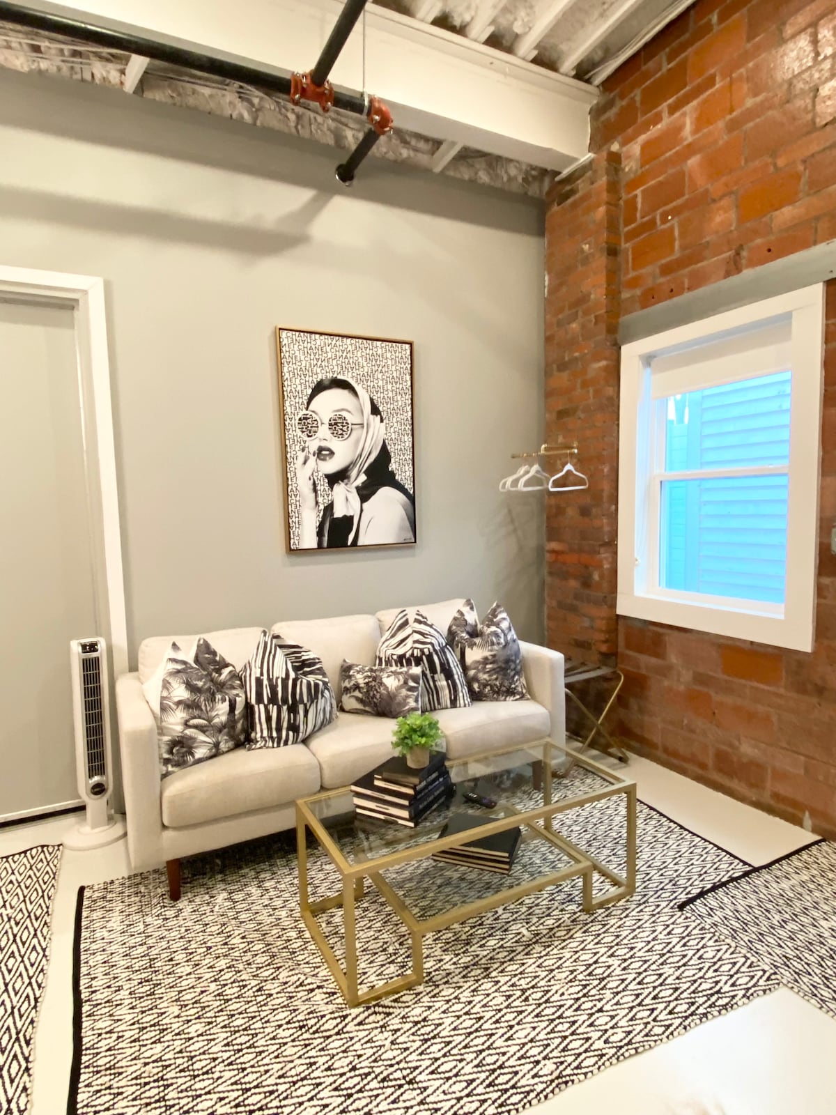 A light-filled seating area features a stylish white sofa adorned with patterned pillows. A glass coffee table rests on an intricate area rug, while a piece of artwork hangs on the wall. Natural light streams through a nearby window, illuminating the brick accent wall.