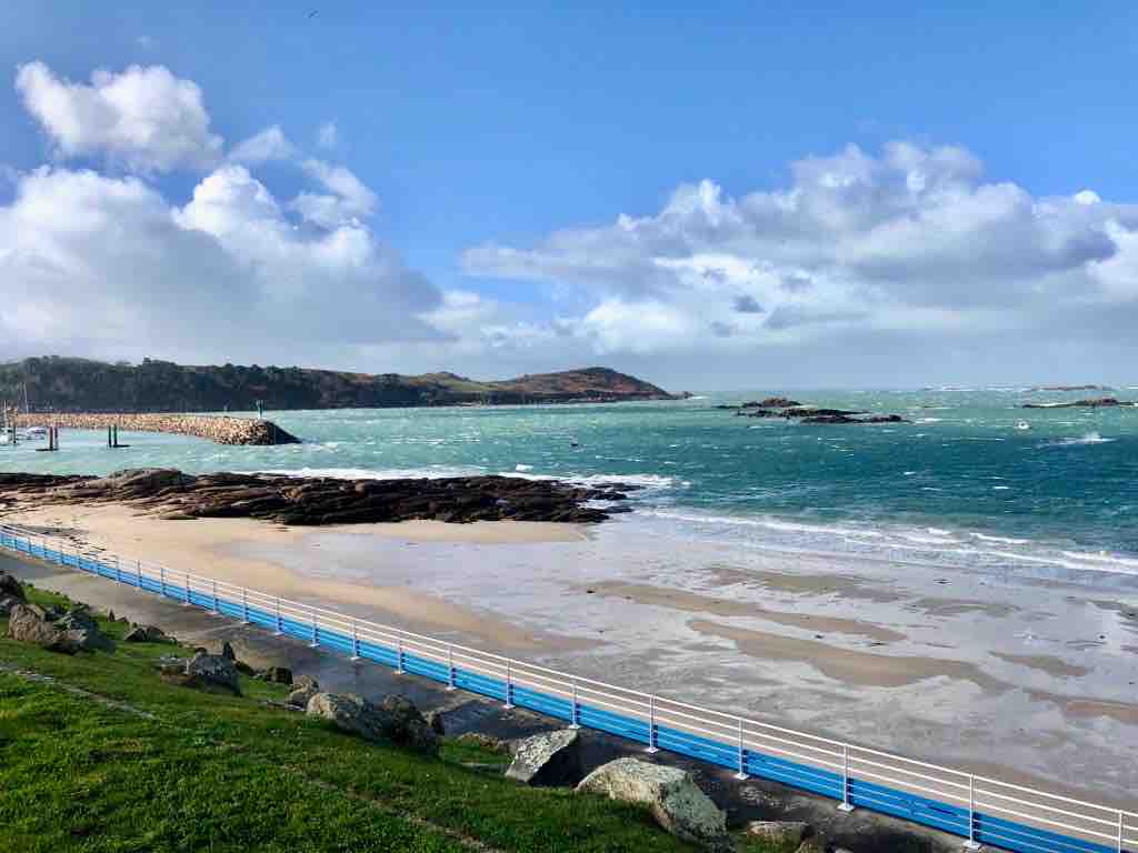A serene beach scene is presented, featuring gentle waves lapping against the sandy shore. A grassy area leads to a walkway outlined in blue. In the distance, rocky formations and hills are visible under a partly cloudy sky.
