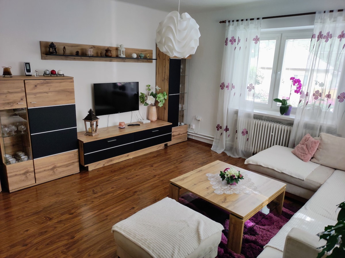A comfortable living area is presented with a light-colored sectional sofa and a wooden coffee table adorned with a floral arrangement. A TV is mounted on a wooden cabinet, while soft curtains with floral patterns frame a window that allows natural light to fill the space.
