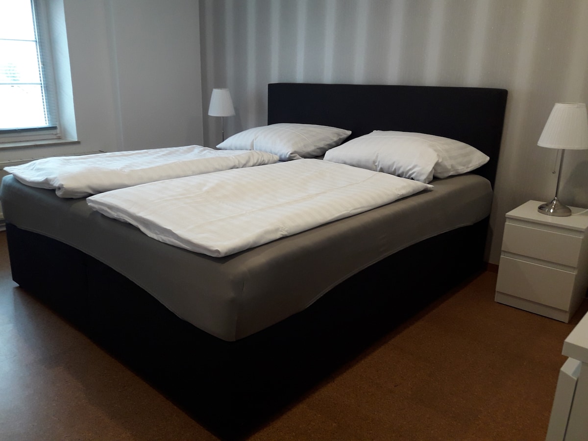 A spacious bedroom features a dark-colored bed with separate mattresses and crisp white bedding. Two bedside lamps are placed on white nightstands. Natural light enters through a nearby window, and the walls are adorned with a subtle striped pattern.