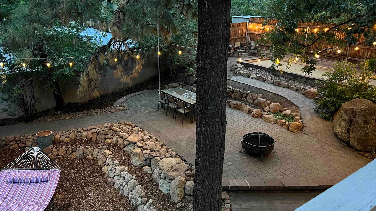 A spacious outdoor area features a meandering stone pathway surrounded by greenery. A long dining table is positioned under string lights, and a firepit is located nearby. A hammock rests to the side, inviting relaxation amidst the natural setting.