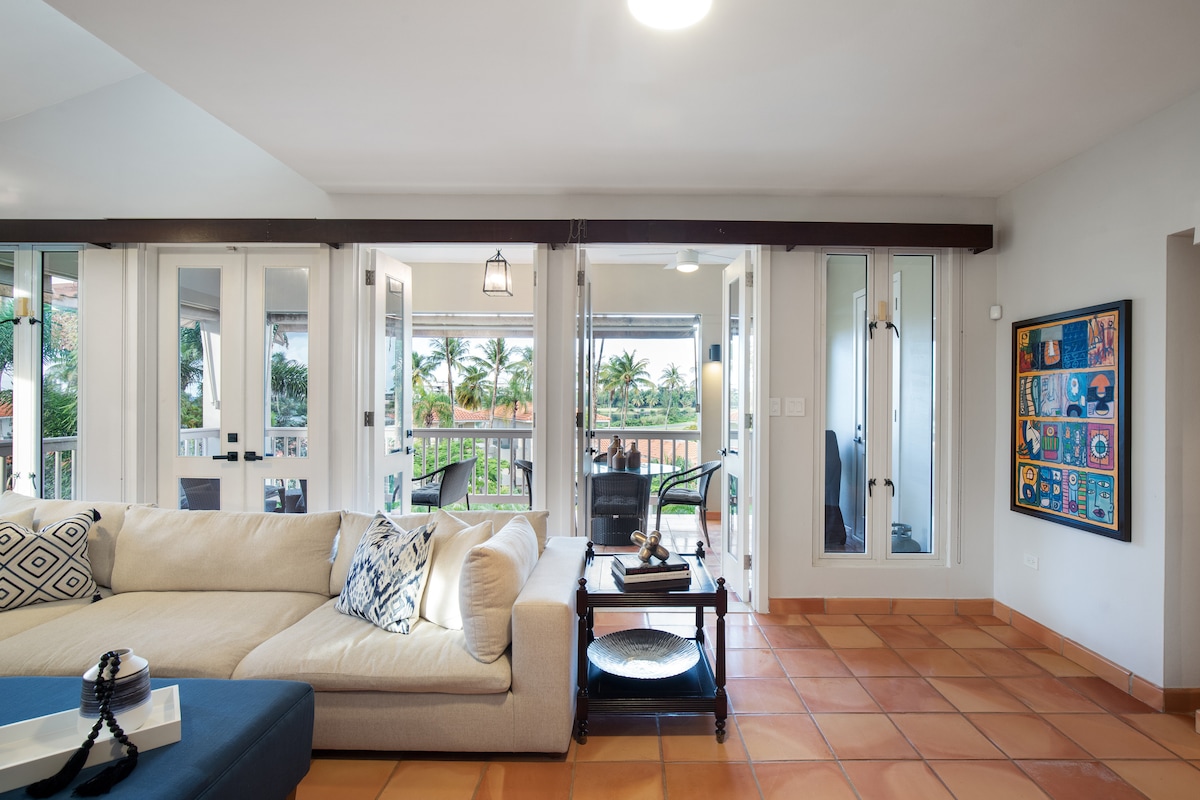 A bright living area features a comfortable sofa adorned with decorative pillows. Large glass doors provide access to the outdoor terrace, allowing natural light to fill the space. A coffee table is positioned in front of the sofa, complemented by a gallery wall of colorful artwork.