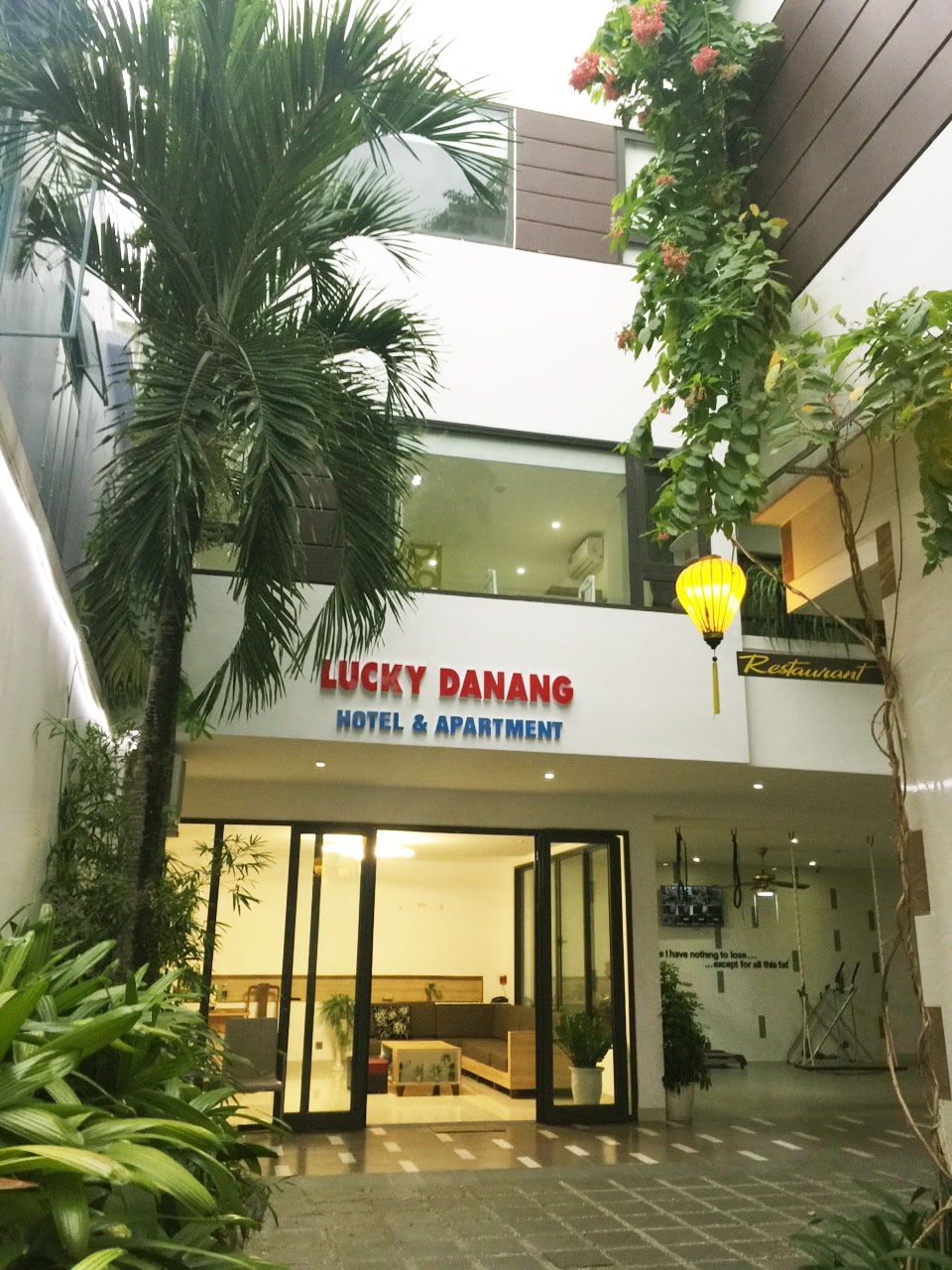 The entrance of Lucky Danang Hotel & Apartment is framed by lush greenery and tropical plants. A welcoming sign displays the hotel's name, and glass doors lead into a bright interior space. A warm ambiance is created by soft lighting and decorative elements.