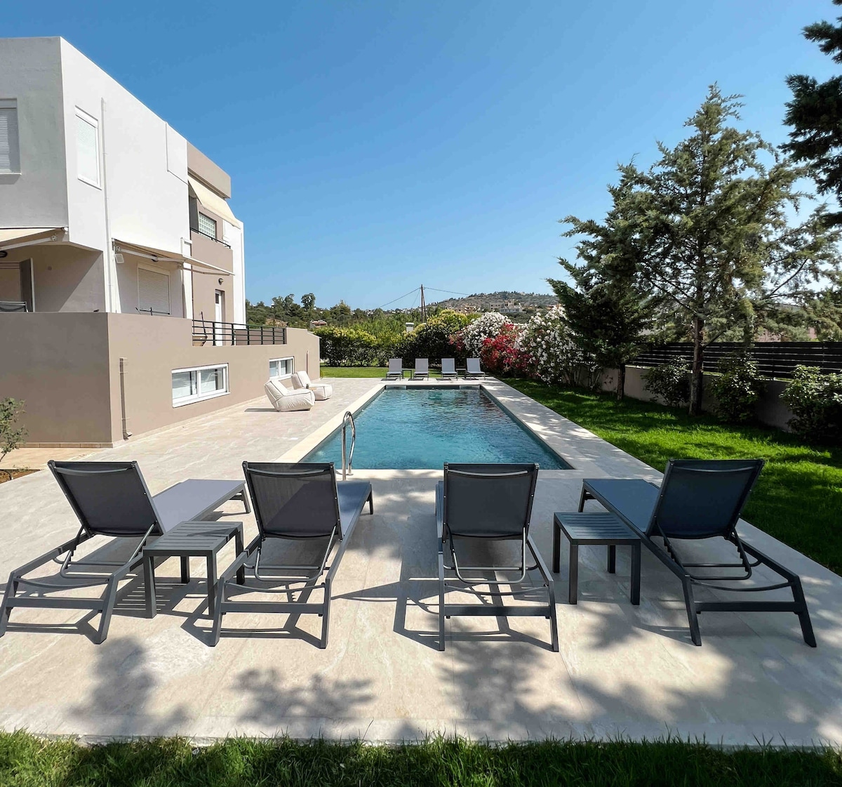 An outdoor area features a private swimming pool surrounded by a spacious patio. Several lounge chairs are positioned neatly beside the pool, offering a relaxing space for sunbathing. Lush greenery and blooming flowers are visible in the background, enhancing the tranquil setting.