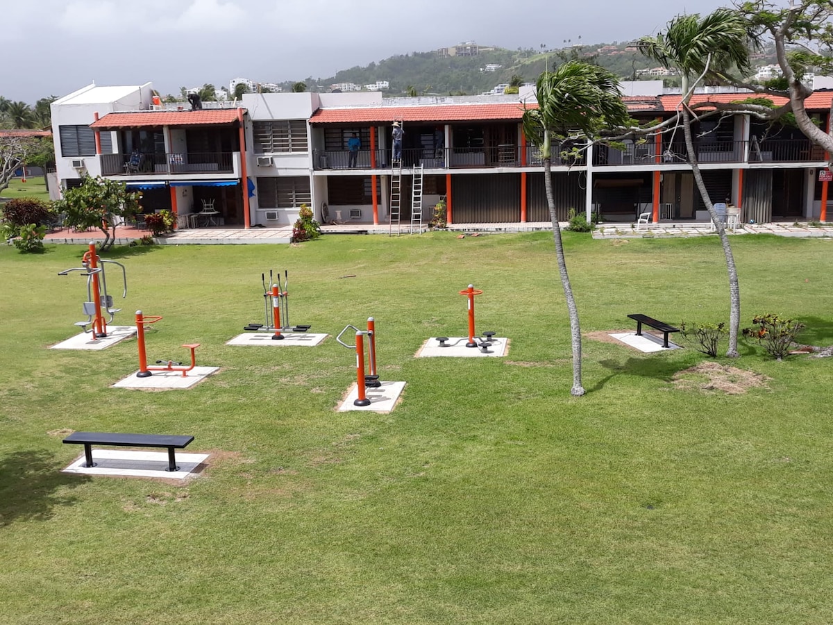 An outdoor area is shown featuring exercise machines set on a grassy lawn, designed for outdoor fitness. The machines have bright orange accents. Behind, a row of condominiums is visible with multiple doors and balconies, surrounded by trees swaying in the breeze.