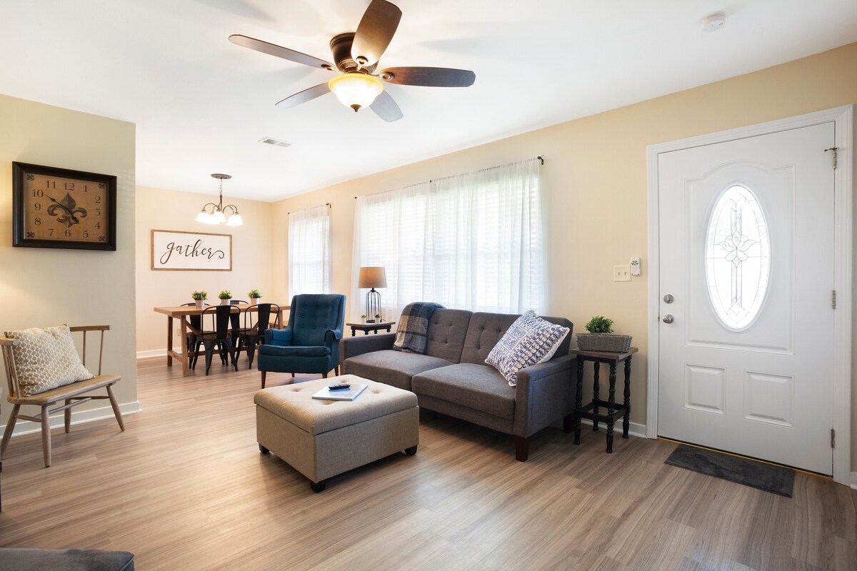A bright living room features a comfortable gray sofa and an ottoman, arranged to invite relaxation. Natural light fills the space through large windows adorned with light curtains. A dining area with a table and chairs is visible in the background, creating an inviting atmosphere.