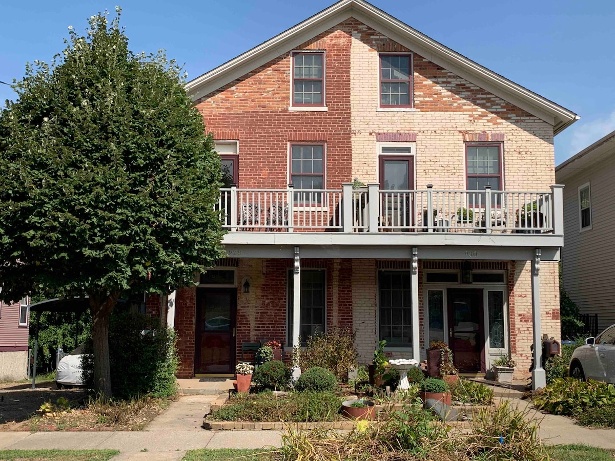 The historic brick building features a two-story facade with a combination of red and cream bricks. A welcoming porch with railing lines the front, adorned with potted plants. Lush greenery surrounds the entrance, creating a pleasant view from the street.