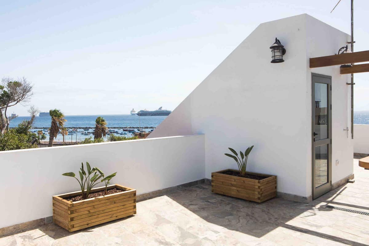 A spacious terrace is displayed, featuring two wooden planter boxes with greenery. In the background, the sea and sandy beach are visible under clear skies, enhancing the outdoor space's atmosphere. The terrace is partially covered, allowing for shaded relaxation.