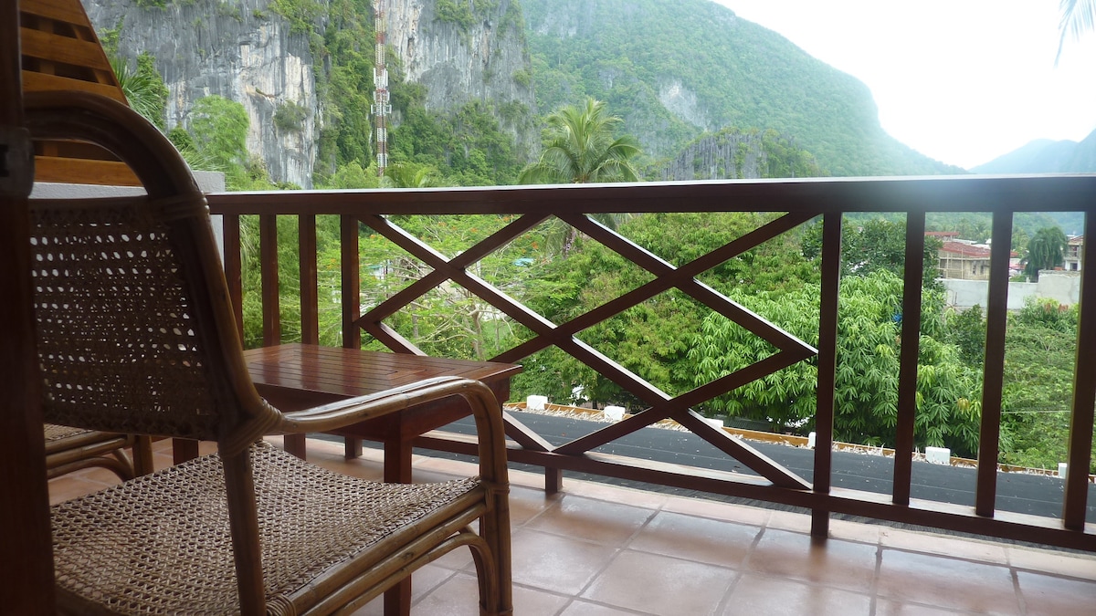 A private balcony displays a wooden railing with an intricate design, overlooking lush greenery and distant hills. A small table and a single chair are positioned on the tiled floor, inviting relaxation while enjoying the scenic views of El Nido's landscape.