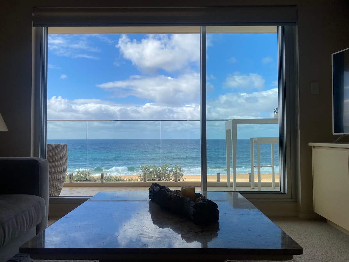 A large glass sliding door offers a clear view of the beachfront and ocean. Soft waves can be seen lapping at the shore under a partly cloudy sky. A stylish coffee table with a decorative centerpiece sits in the foreground, contrasting with the serene outdoor scenery.