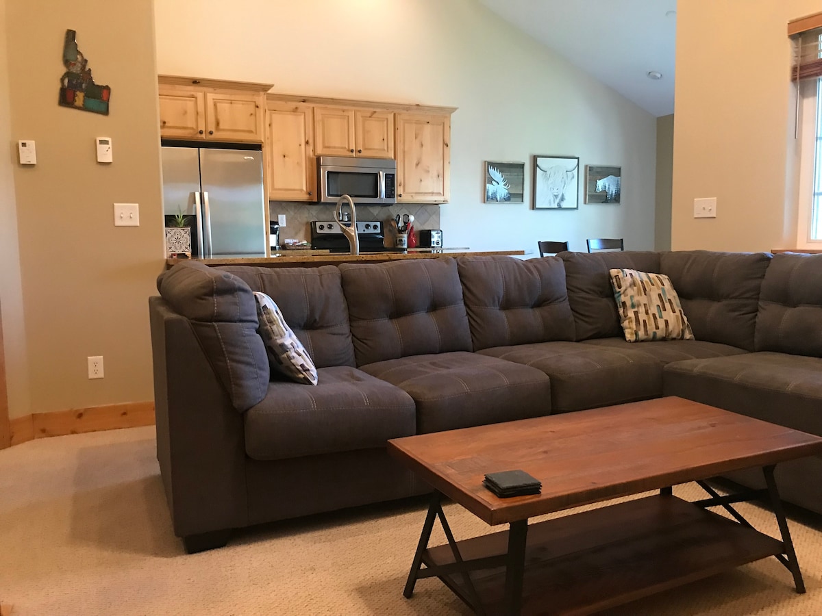 A spacious family room is highlighted by a large sectional sofa with patterned cushions, facing a wooden coffee table. The kitchen area features wooden cabinetry and stainless-steel appliances. Natural light enters through a nearby window, illuminating the cozy gathering space.