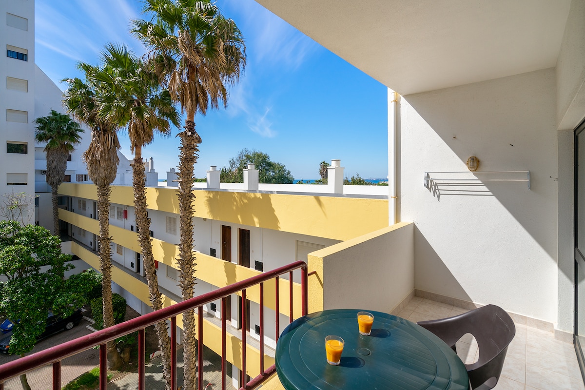 A balcony is depicted with a round table and two glasses filled with a yellow beverage. Palm trees are visible in the background, alongside a view of the surrounding buildings and blue sky. The light-colored railing complements the white walls of the exterior.