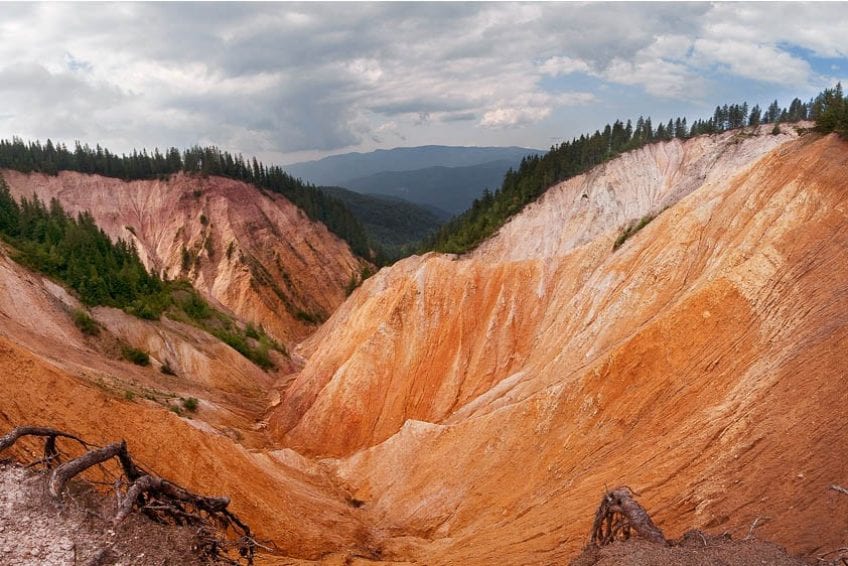 A wide view of a colorful clay ravine displays vibrant shades of red, orange, and cream, contrasted against deep green forested hills. The sky shows an overcast backdrop with soft clouds creeping over the landscape, highlighting the natural beauty of the Apuseni Mountains.