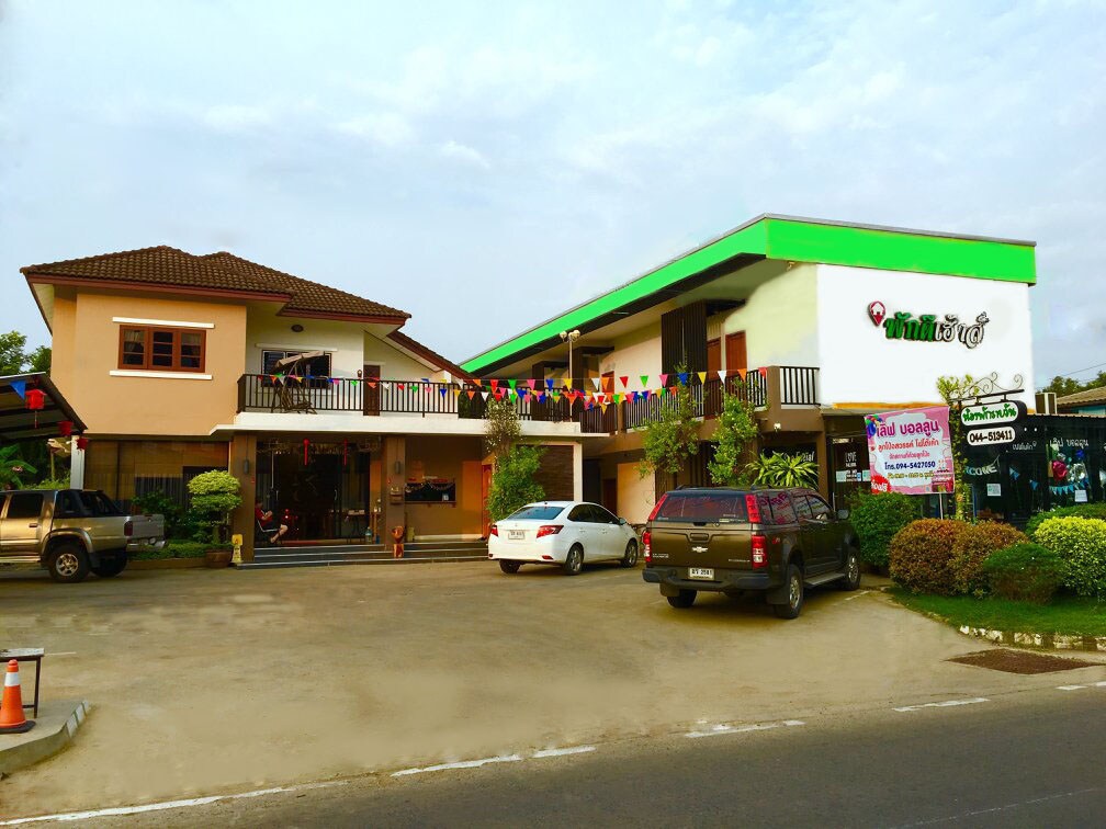 A two-story building features distinct architectural elements, with a sloped roof and a vibrant green overhang. The front is landscaped with tidy shrubs and decorative flags. A spacious parking area is visible, accommodating several vehicles, providing easy access to the entrance.