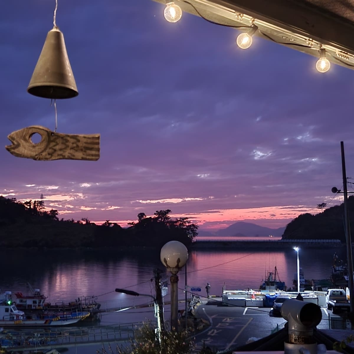 A serene seascape is captured at sunset, with vibrant hues reflecting on calm waters. Fishing boats are lined up in the harbor, while decorative elements like a wooden fish and hanging lights add charm to the scene. The distant landscape includes gentle hills under a colorful sky.