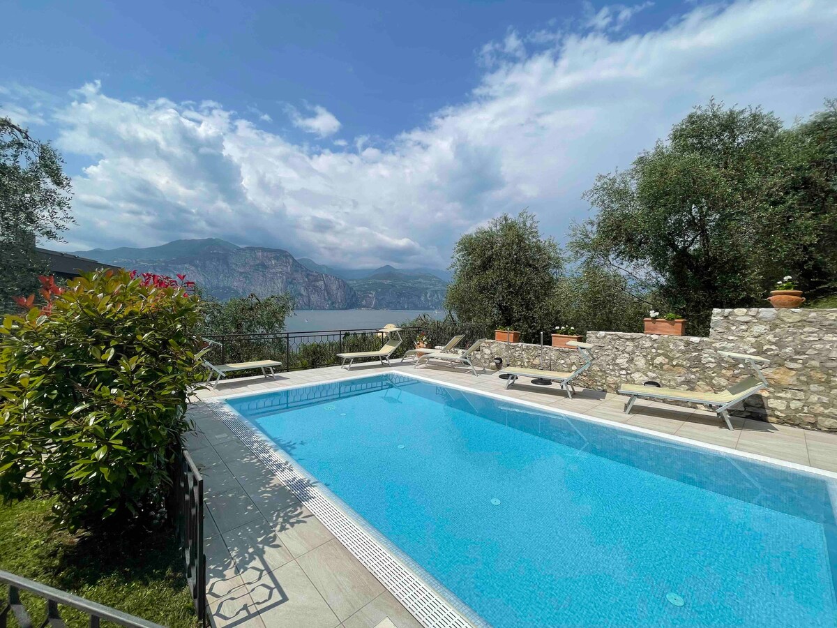 A sparkling swimming pool surrounded by greenery is presented in the foreground, with lounge chairs positioned along the deck. The backdrop features a scenic view of rolling hills and distant mountains, complemented by flowering plants and stone walls that define the area.