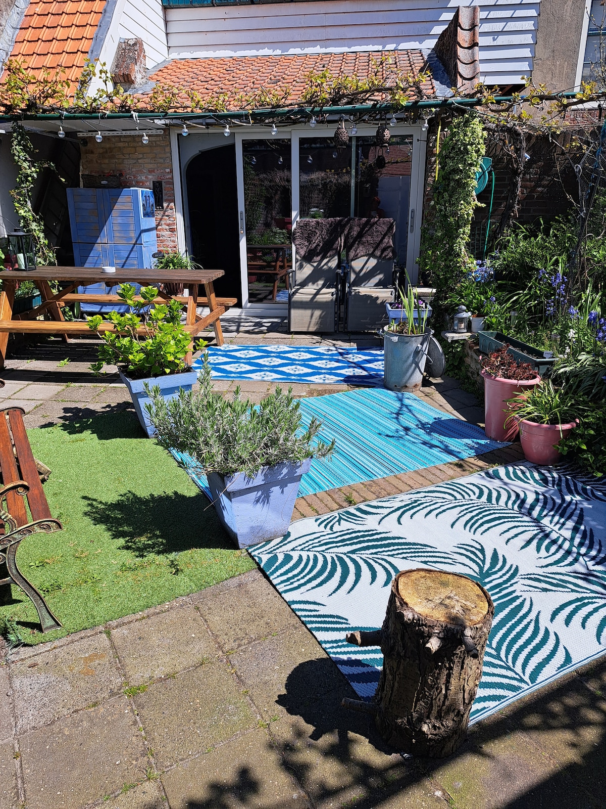 A private garden is displayed, featuring patterned rugs and a mix of outdoor seating options. Potted plants and greenery add a natural touch. A wooden table is positioned nearby, creating a cozy area for relaxation. The entrance to the apartment is visible in the background.