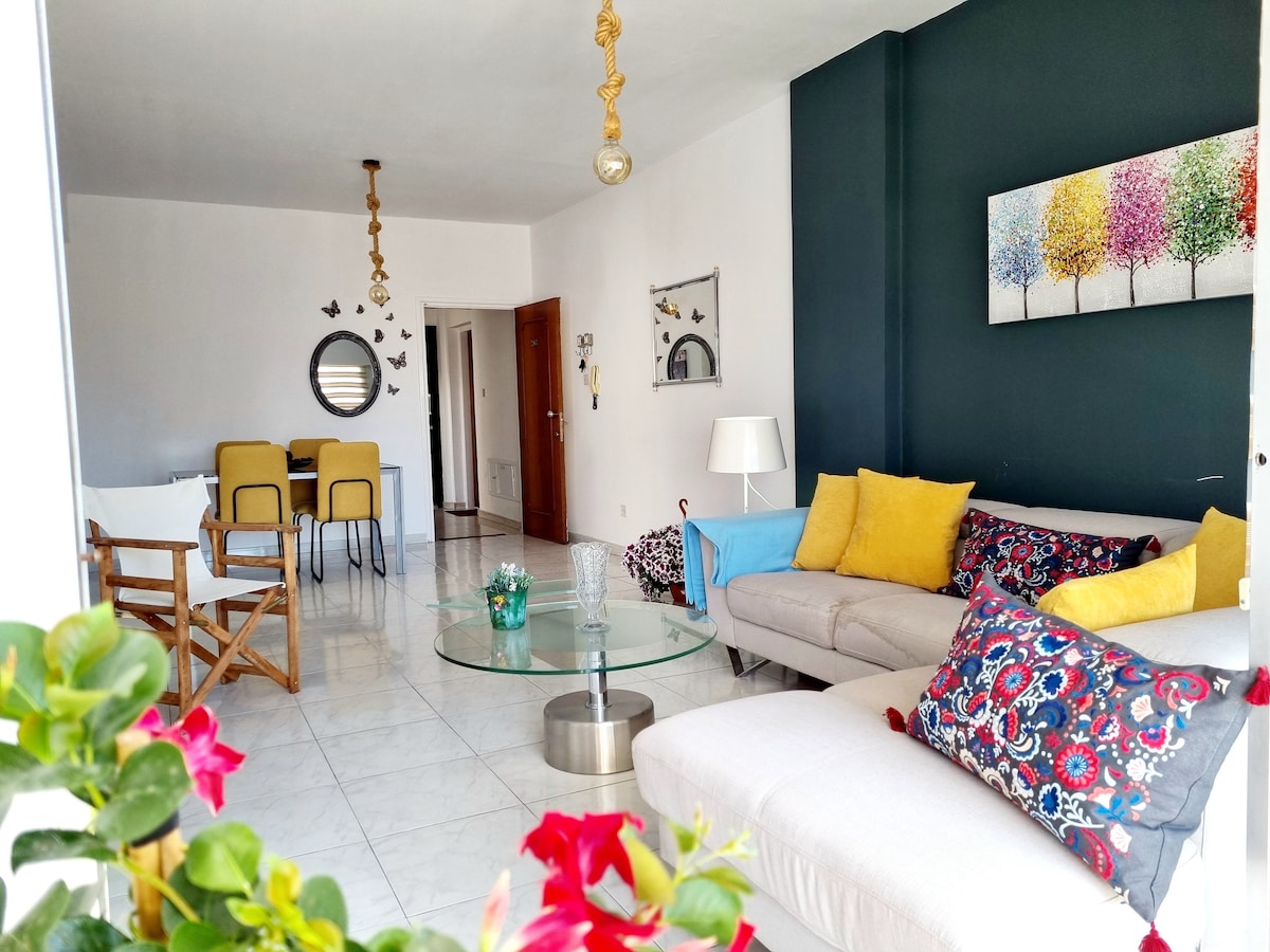 A bright living area features a light-colored sectional sofa adorned with patterned cushions. A round glass coffee table occupies the center space, surrounded by greenery. The walls are partially painted in a deep blue tone, complemented by colorful artwork and an inviting dining setup in the background.