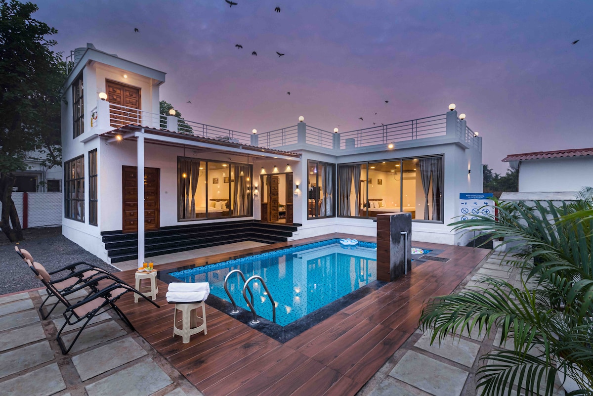 The villa features a private swimming pool surrounded by a wooden deck and lounge chairs. Large glass doors open into spacious living areas, with outdoor staircases leading to an upper terrace. The warm evening sky is illuminated by surrounding lights, with birds flying overhead.