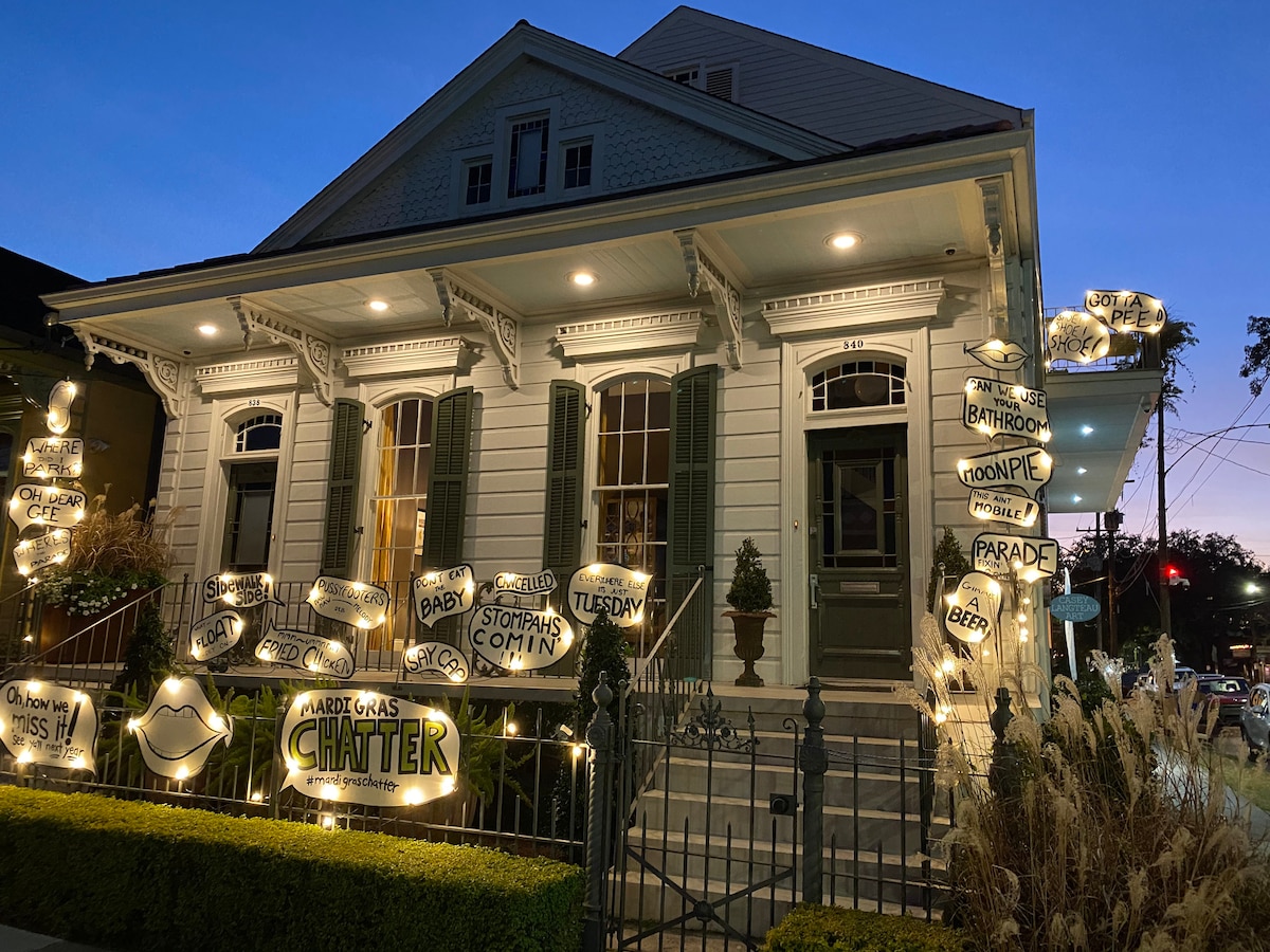A historic building is adorned with illuminated signs featuring playful phrases and mouth shapes. The white exterior contrasts with green shutters. Soft lighting enhances the welcoming front porch, surrounded by manicured landscaping and wrought iron fencing under a twilight sky.