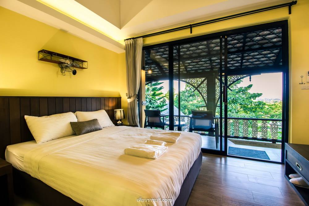 A spacious room features a large bed with crisp linens, complemented by a seating area next to a sliding glass door. The door opens to a balcony with scenic views of greenery, while warm yellow walls provide a cozy atmosphere.