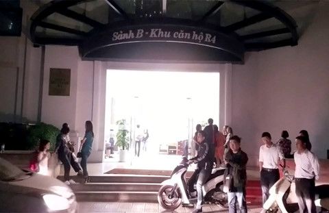 An entrance to a building is shown, featuring a covered pathway marked with a sign. Groups of people are gathered outside, with a motorcycle parked nearby. The bright interior lights create a welcoming contrast against the evening ambiance.