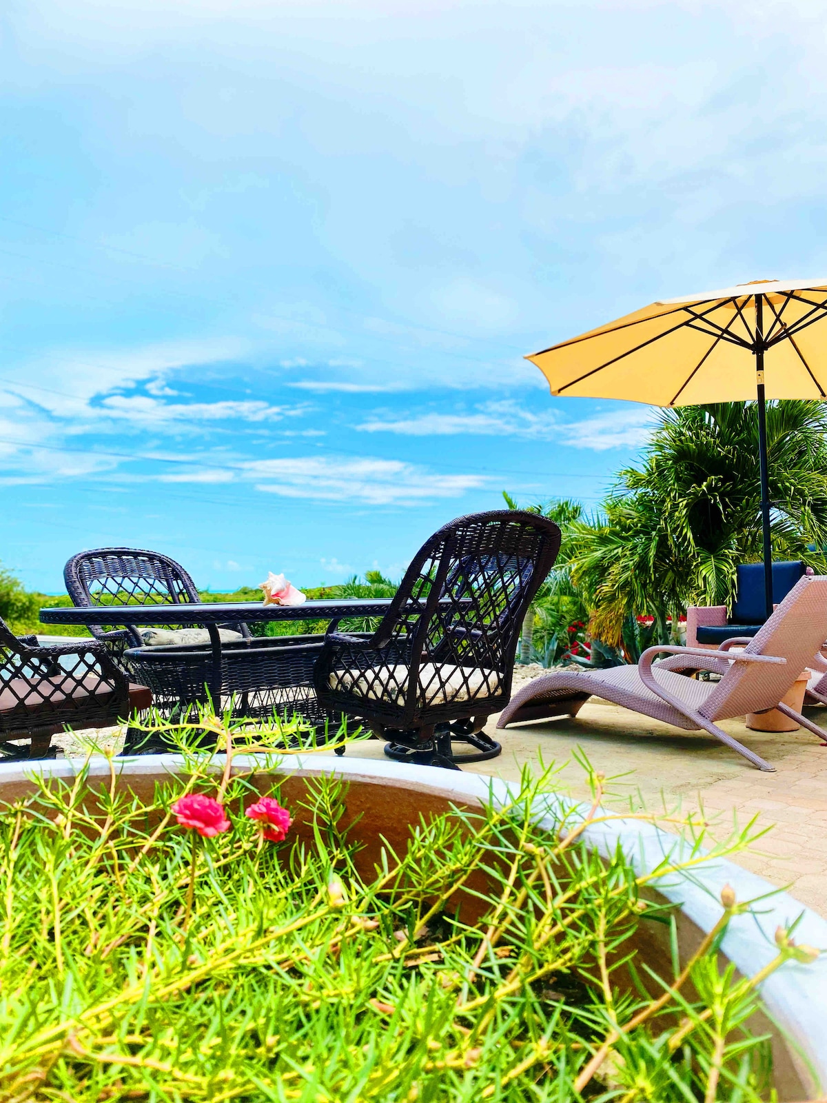 An outdoor seating area is featured with woven chairs and a round table, set against a clear blue sky. A large yellow umbrella provides shade, while potted greenery with pink flowers adds a natural touch to the serene environment.